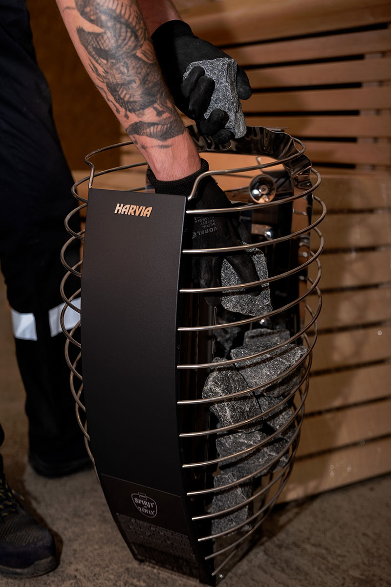 Närbild av korgen till en HARVIA elektrisk spis: mörkgrå bastustenar placeras i svarta handskar.